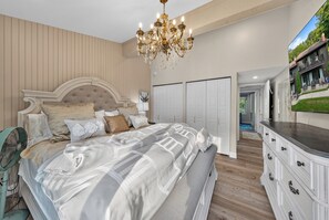 Een slaapkamer met een groot bed, een kroonluchter, een kaptafel en uitzicht op een huis door een raam.
