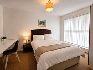 Ein Hotelzimmer mit Bett, Nachttisch, Lampe, Stuhl und einem Fenster mit Jalousien.