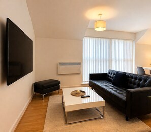 Een moderne woonkamer met een flatscreen televisie aan de wand, een zwarte leren bank, een witte salontafel en een groot raam met jaloezieën.