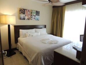 A hotel room with a bed, a bedside table, a lamp, a wall art piece, and a window with curtains. - Spacious Dawn Beach 3-bedroom condo in delightful Upper Prince's Quarter (Upper Prince's Quarter)