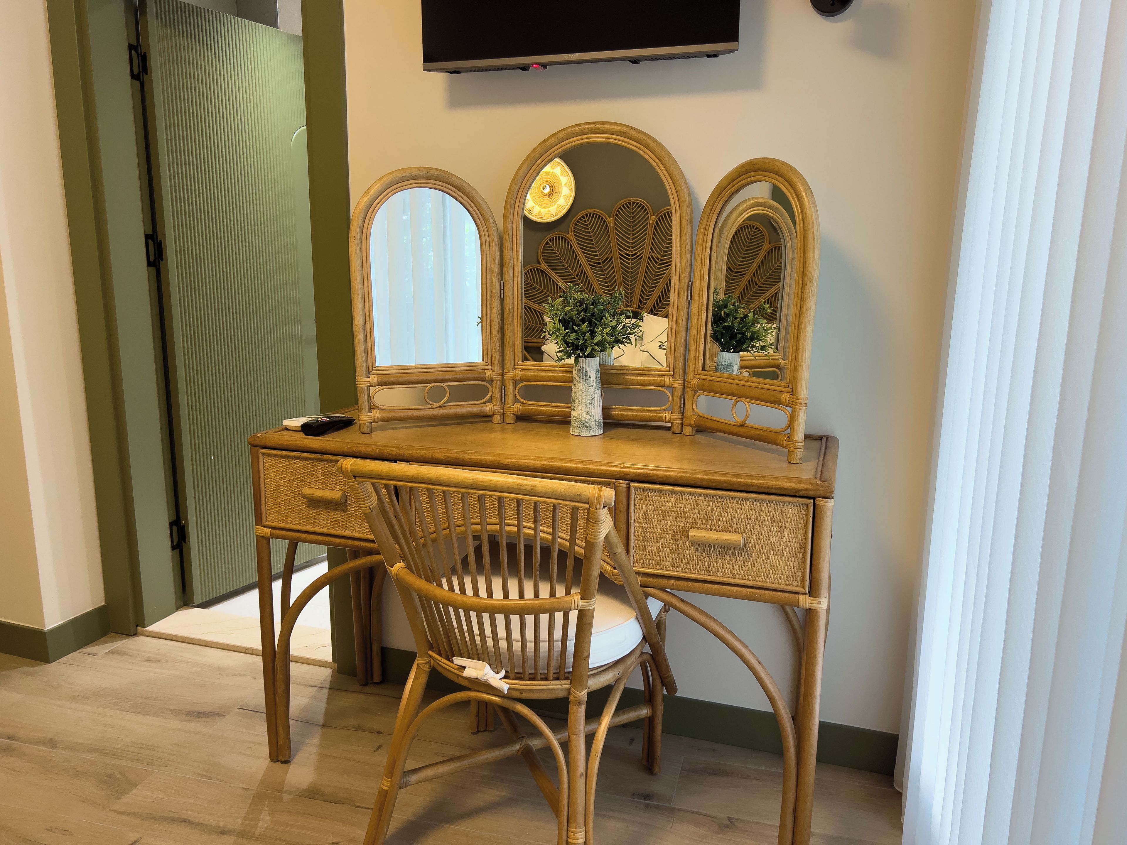 Une pièce comprenant une coiffeuse en bois, trois miroirs, une chaise en osier et une plante en pot.