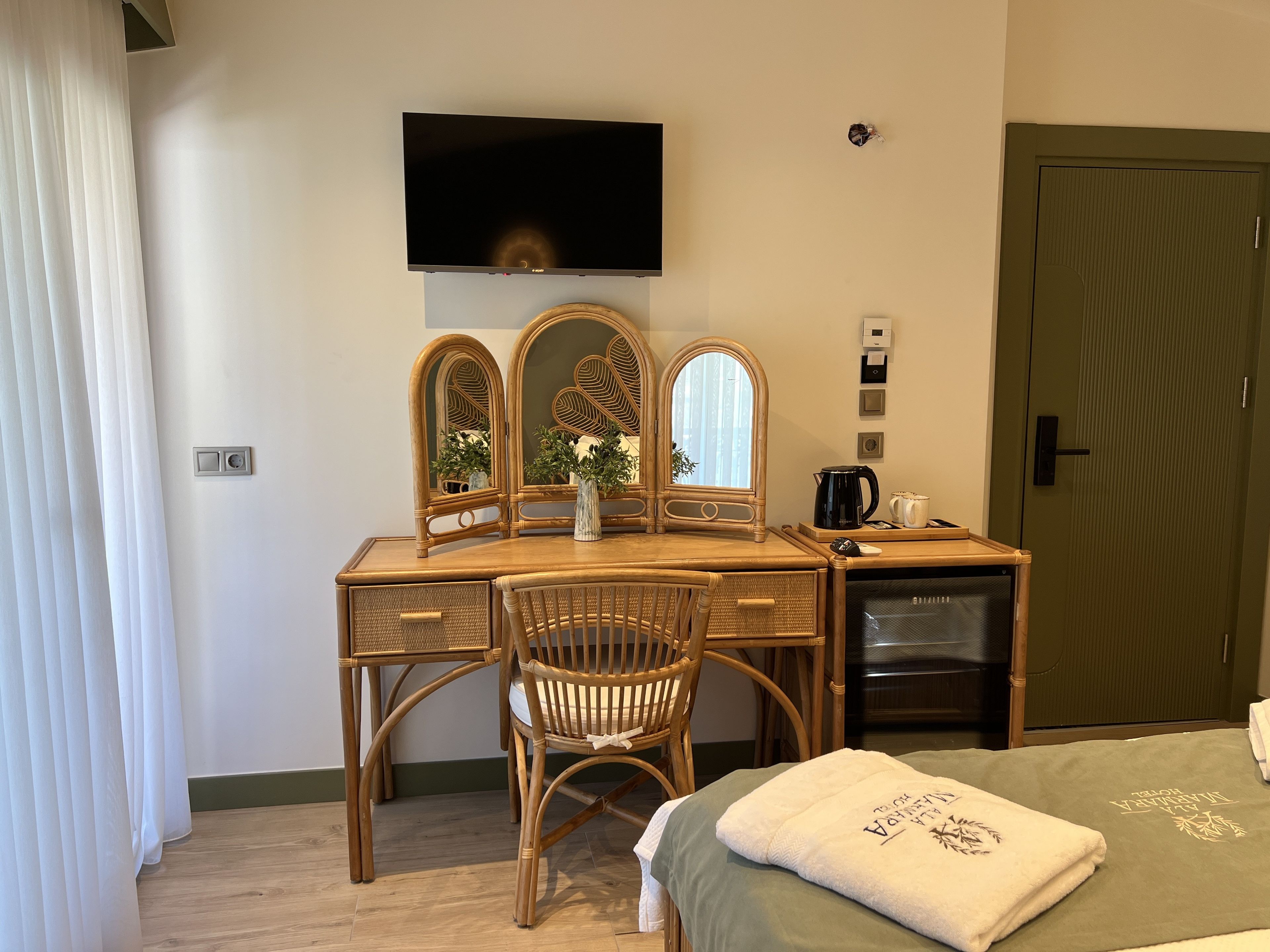 Habitació d'hotel amb escriptori de fusta, cadira, televisió penjada a la paret, cafetera i llit amb estri de color verd i tovallola amb text.