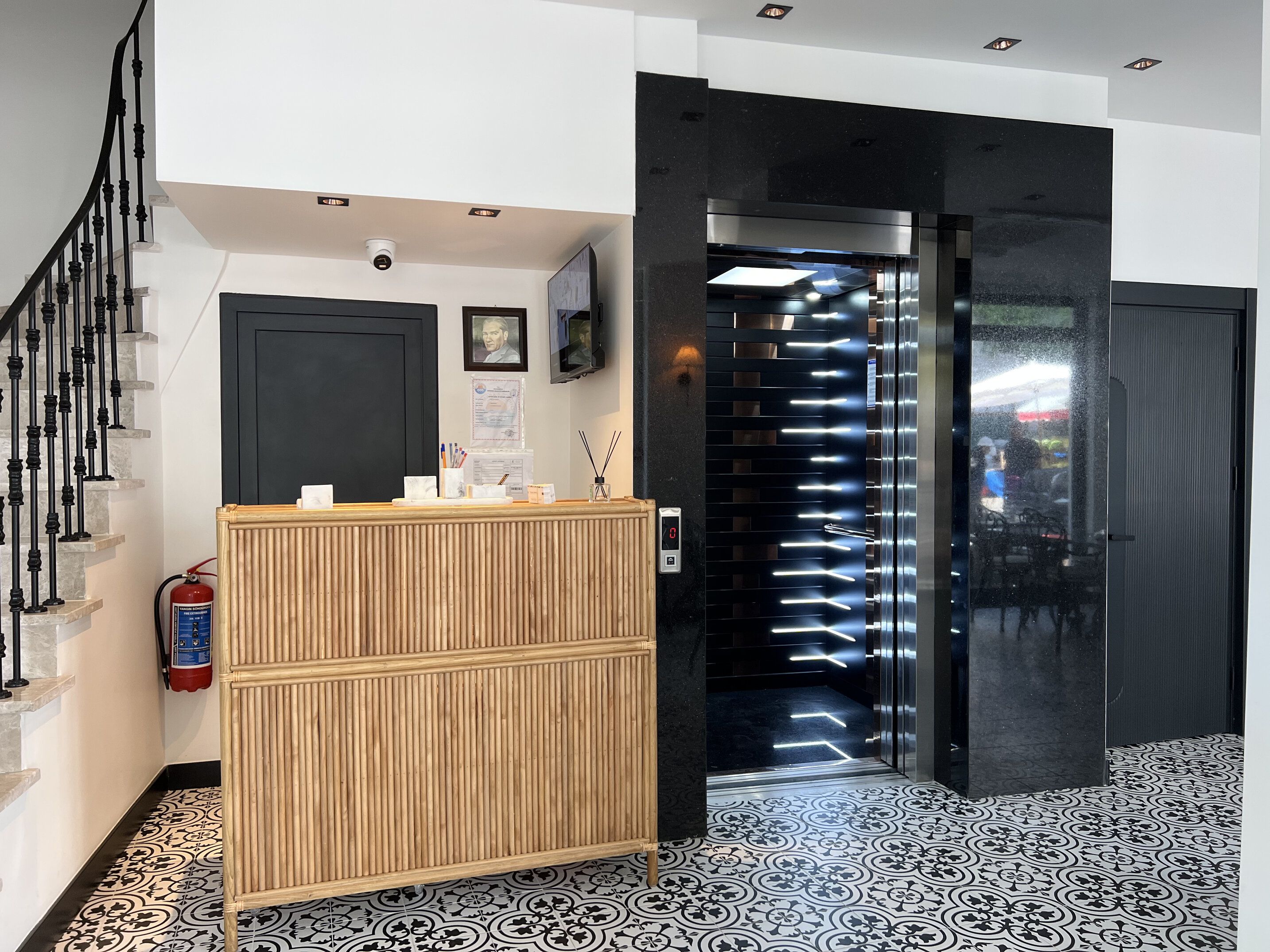 A reception area with a wooden counter, a fire extinguisher, a digital display, and a staircase.