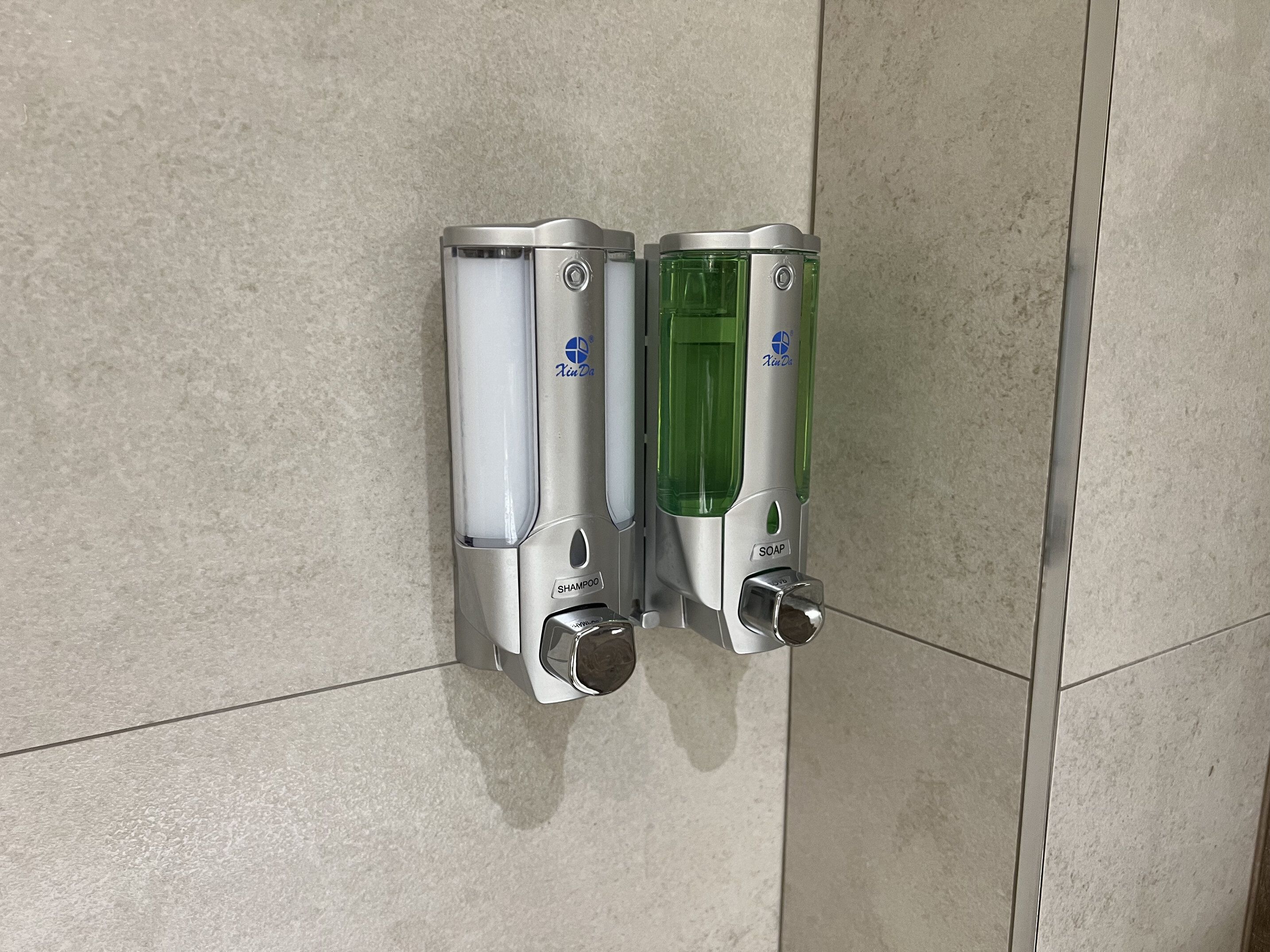 Baño con dos dispensadores de jabón para manos fijos en la pared, uno con líquido transparente y el otro con líquido verde.