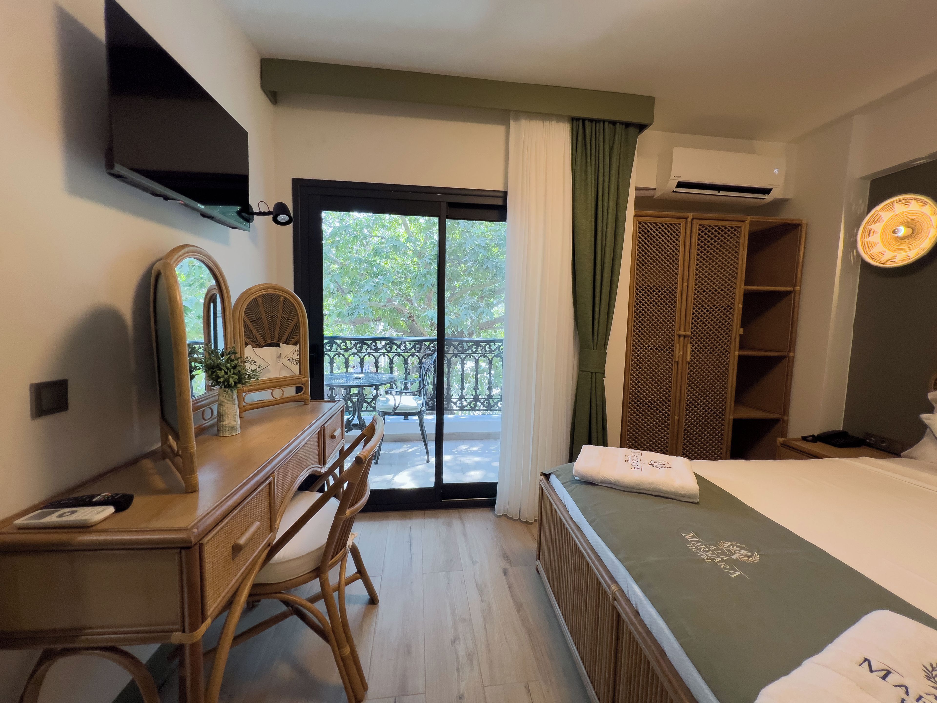 Una habitación de hotel con cama, escritorio con espejo, silla, balcón con vista a árboles y armario.
