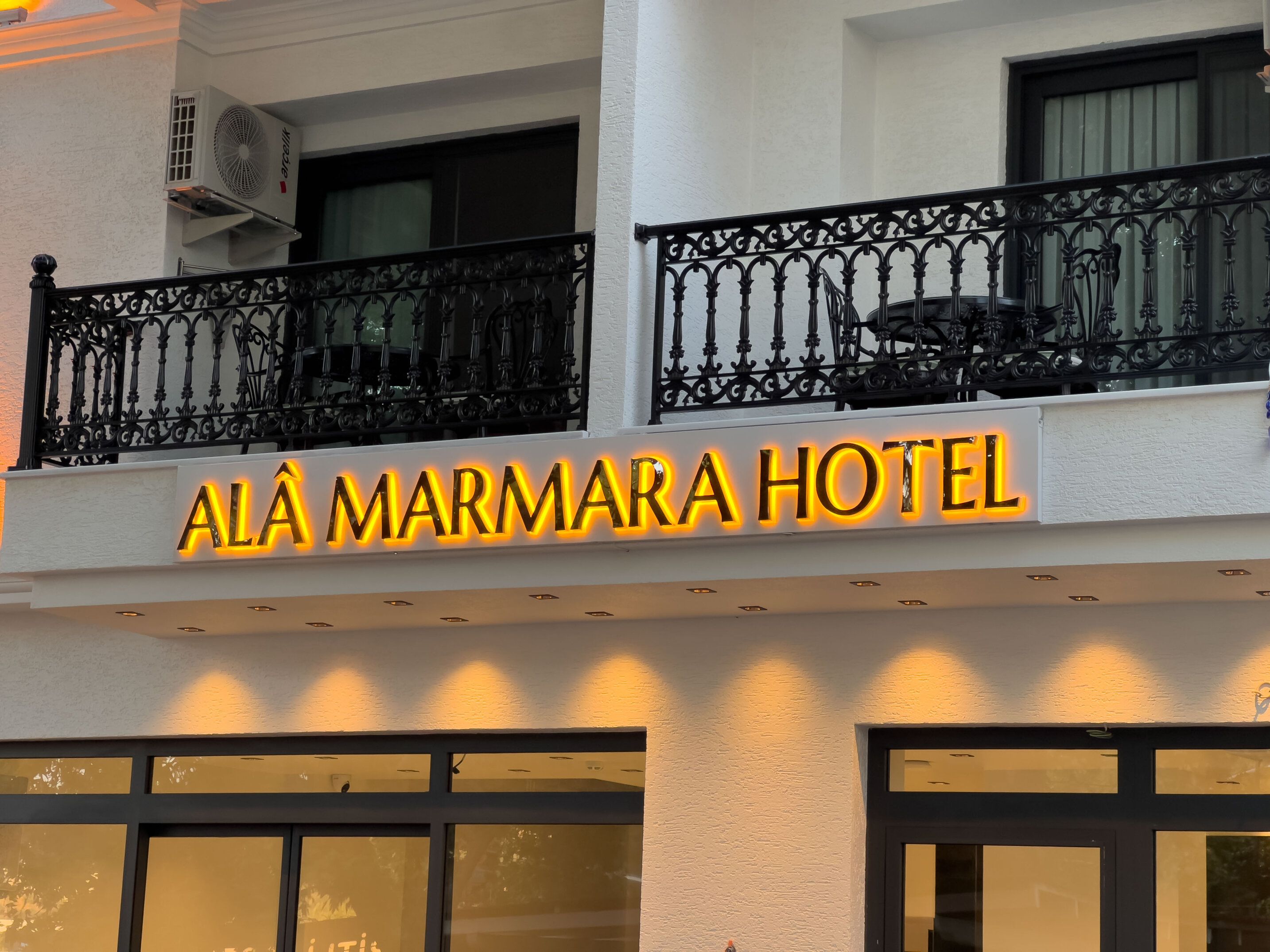 Exterior del Hotel Alá Marmara con un llamativo letrero iluminado, un balcón con barandillas negras ornamentadas y una unidad de aire acondicionado de gran tamaño adosada al edificio.