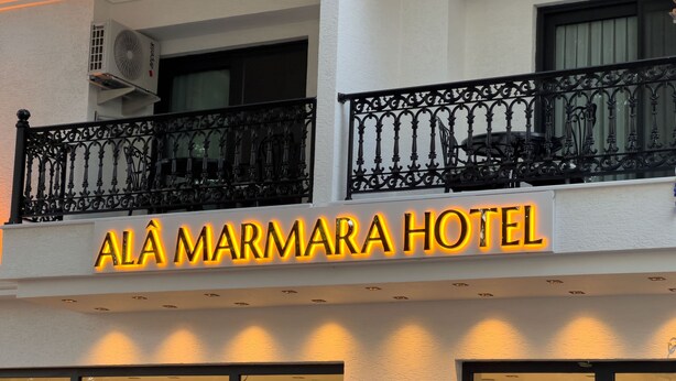 The exterior of Alá Marmara Hotel with a prominent illuminated sign, a balcony with ornate black railings, and a large air conditioning unit attached to the building. - Alâ Marmara Hotel (Marmara)