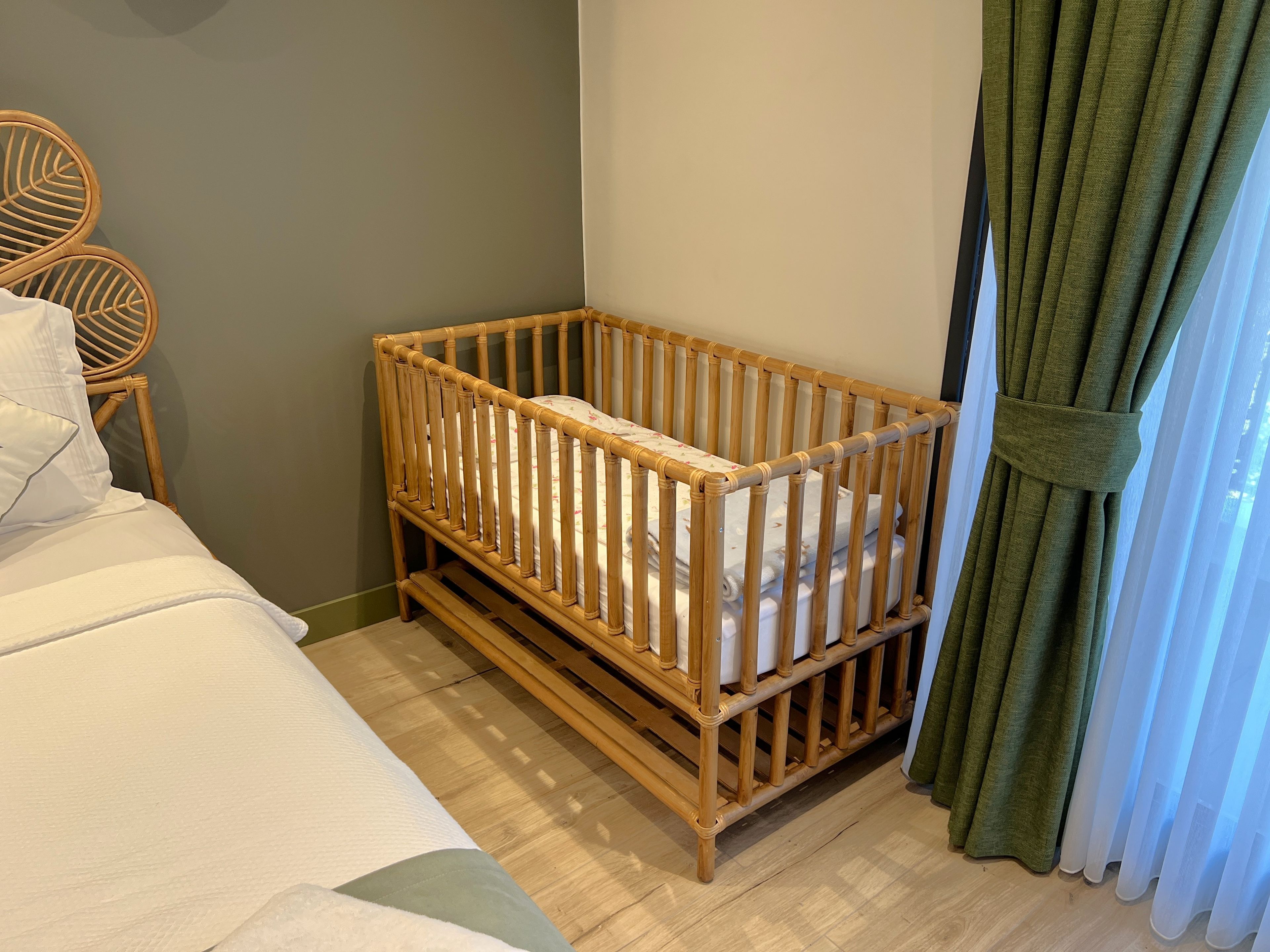 Une pièce avec un berceau en bois, un lit avec une literie blanche, une chaise en osier et des rideaux verts.