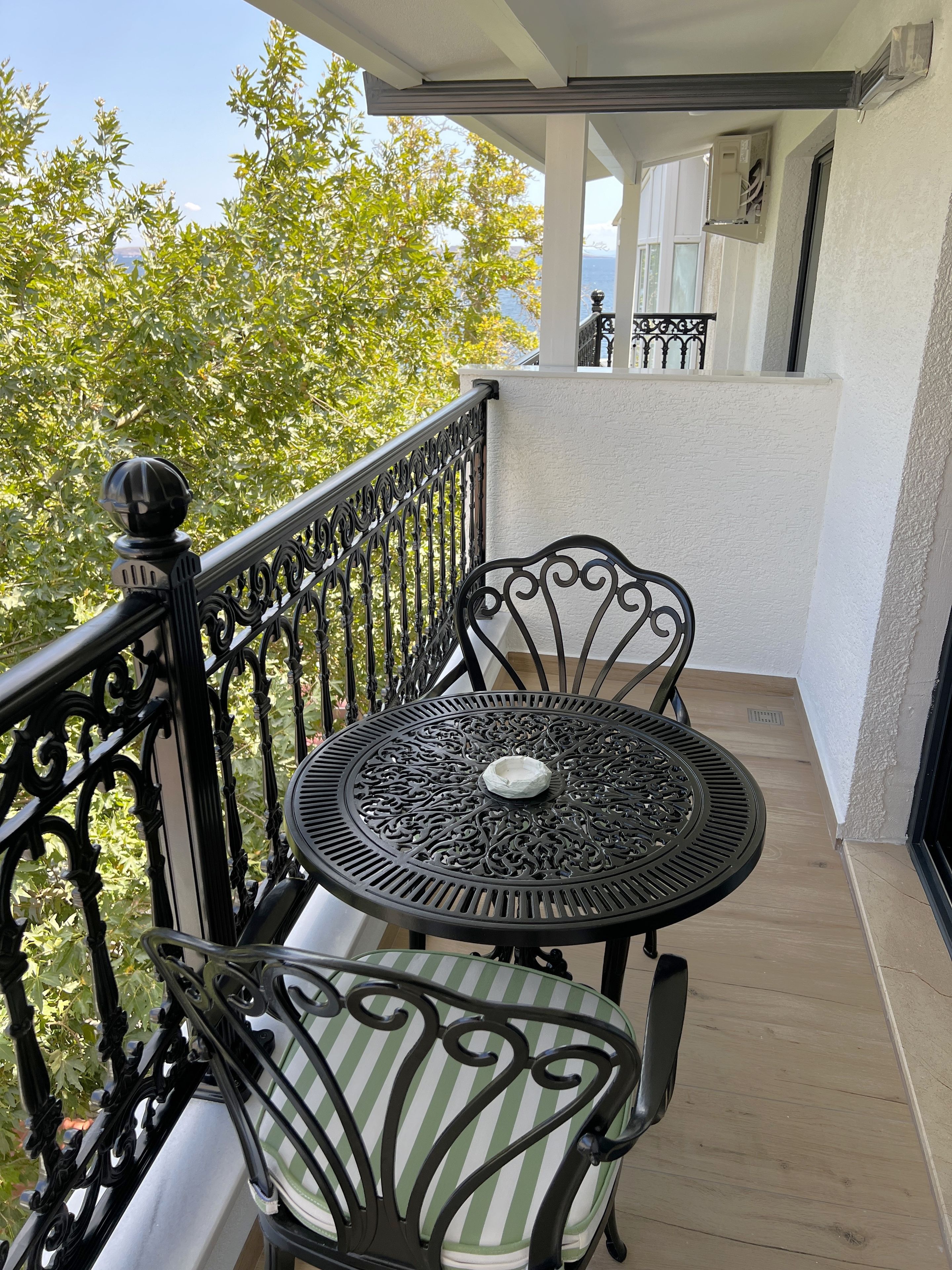 A balcony with a round metal table and two chairs, a railing, and a view of trees and a building in the background.