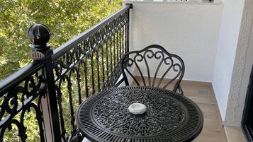 A balcony with a round metal table and two chairs, a railing, and a view of trees and a building in the background.