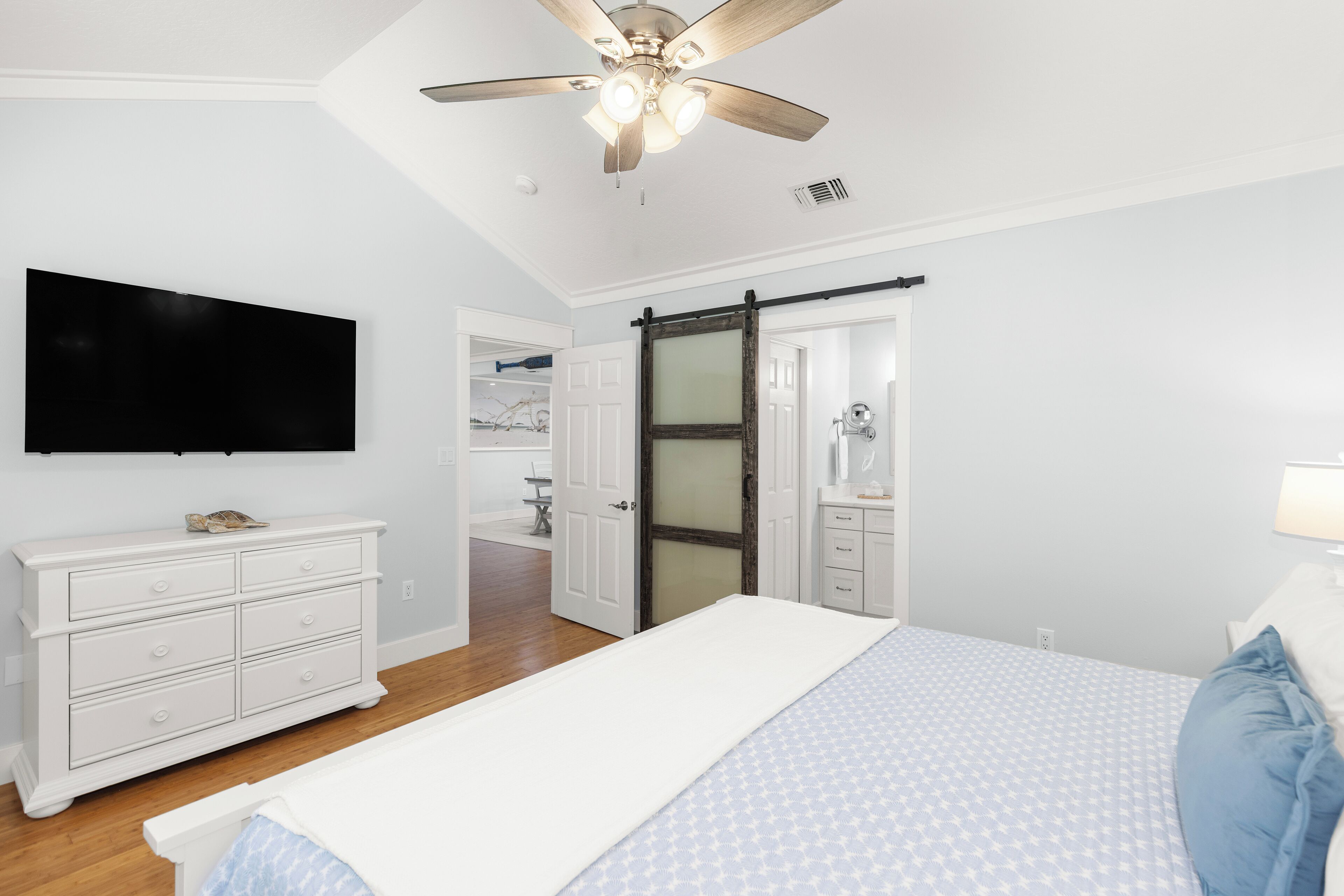 A bedroom with a bed, a dresser, a ceiling fan, a television, and a bathroom visible through an open door.
