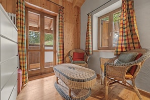 A wooden interior with a wicker ottoman, two wicker chairs, a window with curtains, and a door with a view of greenery outside.