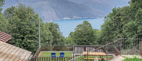 A residential area with a view of a lake and mountains, a fenced yard with chairs, and a small building with a sloped roof.