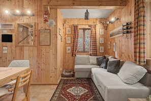 A cozy cabin interior with wooden walls, a sofa, a dining table with chairs, a rug, and various wall decorations including framed pictures and a lantern.