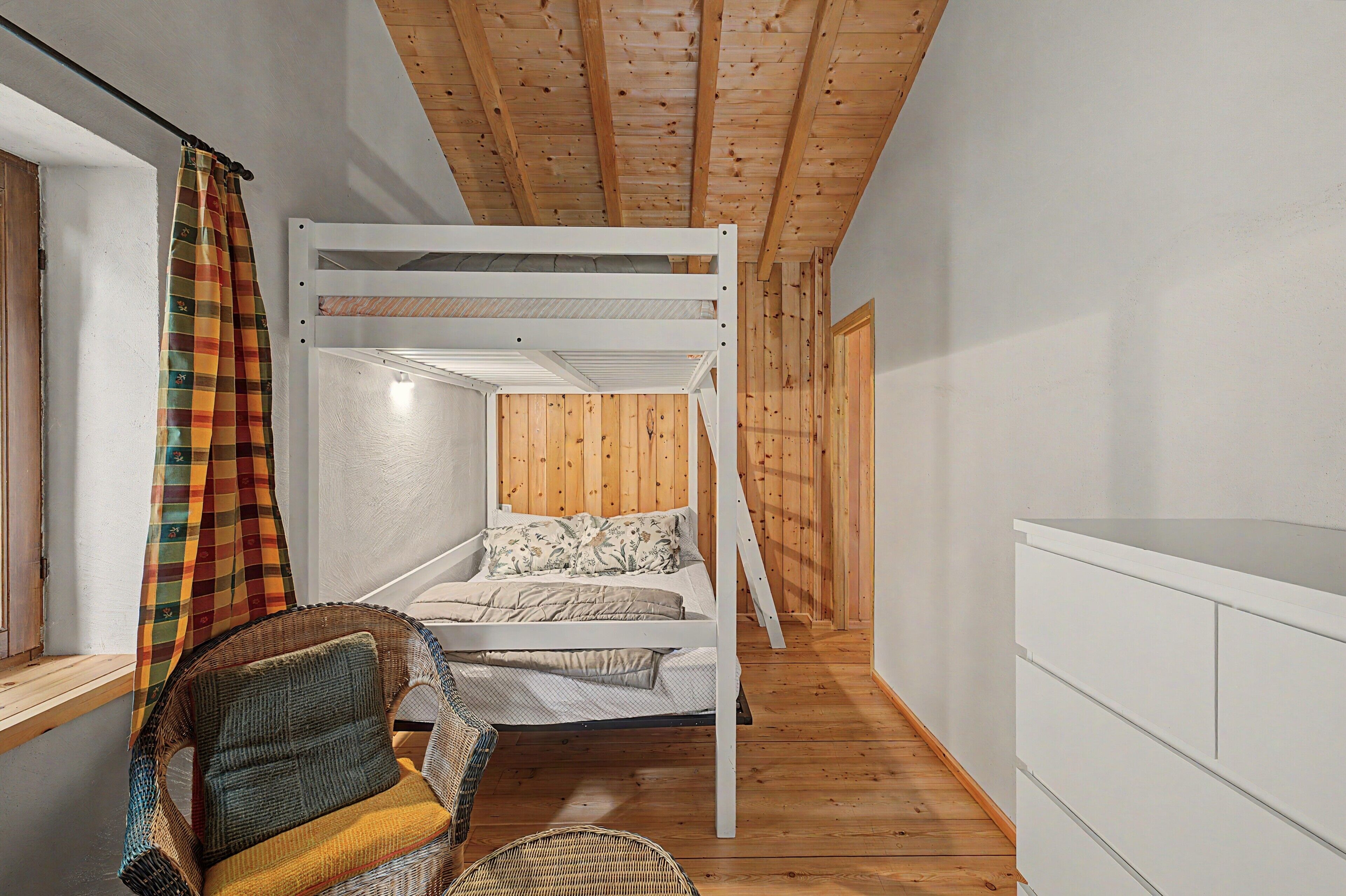 Una cama con literas de estructura blanca, una silla de mimbre y suelo de madera.