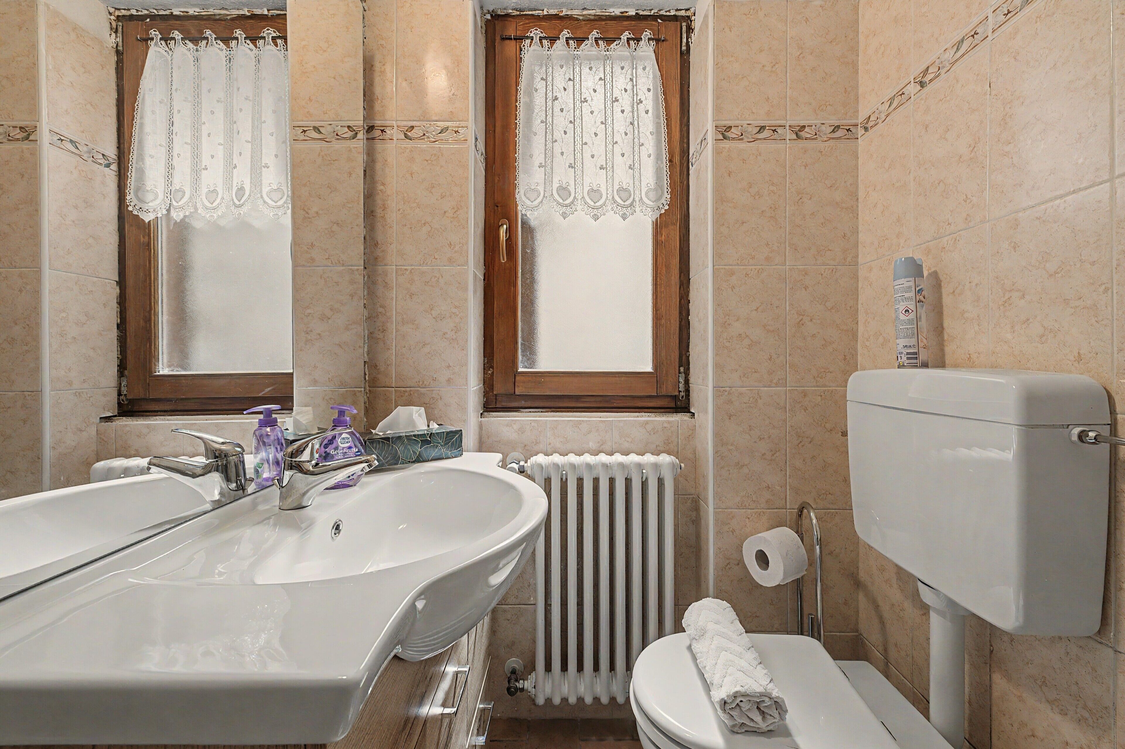Et badeværelse med dobbeltvask, toilet, radiator og et vindue med gardin.