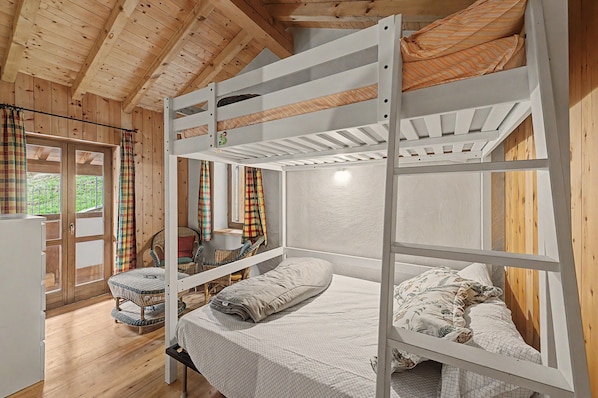 A bunk bed room with a wooden ceiling, a wicker chair, and a window with curtains.