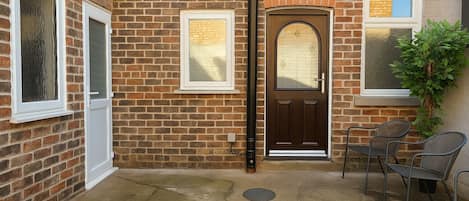 A brick house with a brown door, white-framed windows, and a small patio with chairs.