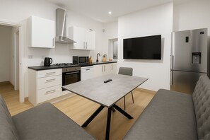 Eine moderne Küche mit weißen Schränken, einer Edelstahl-Dunstabzugshaube, einer Mikrowelle und einem eingebauten Backofen. Es gibt einen grauen Esstisch mit einer Fernbedienung darauf und ein graues Sofa im Wohnbereich.