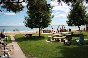 An outdoor seating area with picnic tables and chairs, a swing set, and a view of the beach. - Waterfront 2-Bedroom Villa with Lake Huron Views & Sandy Beach Access (Tawas City)
