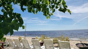Et siddeområde ved stranden med hvide plastikstole og en trillebør, med udsigt over et roligt hav under en klar himmel.