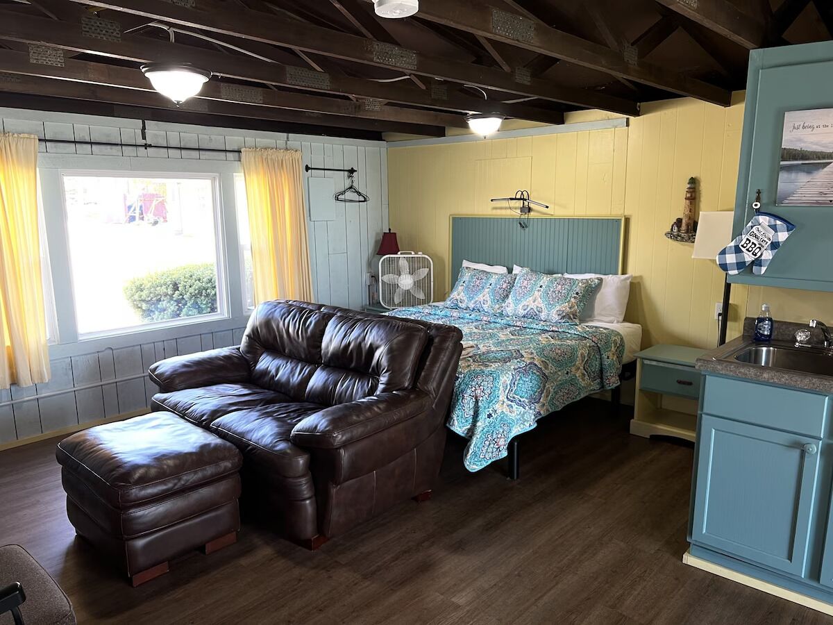 A cozy room with a leather sofa, a bed with patterned bedding, a small kitchenette, and a window with curtains.