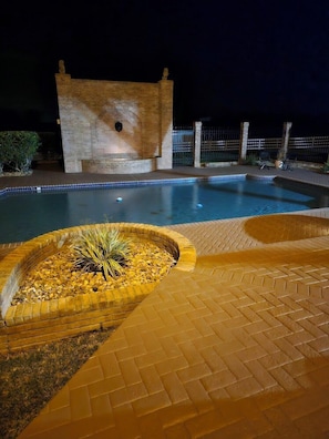 Ein Schwimmbecken mit einer Ziegelstruktur im Hintergrund, umgeben von einer Ziegelterrasse und einer kleinen Gartenanlage mit Pflanzen.