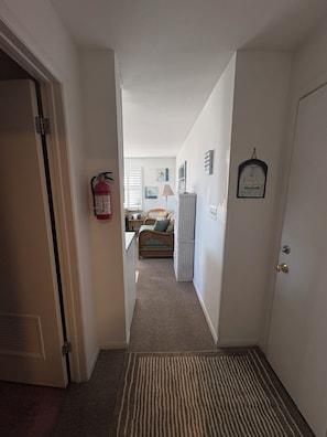 A hallway leading to a bedroom with a bed, a nightstand, and a lamp. There is a fire extinguisher mounted on the wall. - Ocean Views • 1-BR Ocean City NJ Condo • Beach Block • Boardwalk Ramp • King Bed (Ocean City)