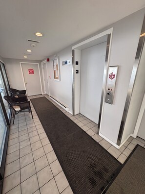 A hotel hallway with a fire pump room sign, an elevator with a control panel, and a chair near the entrance.
