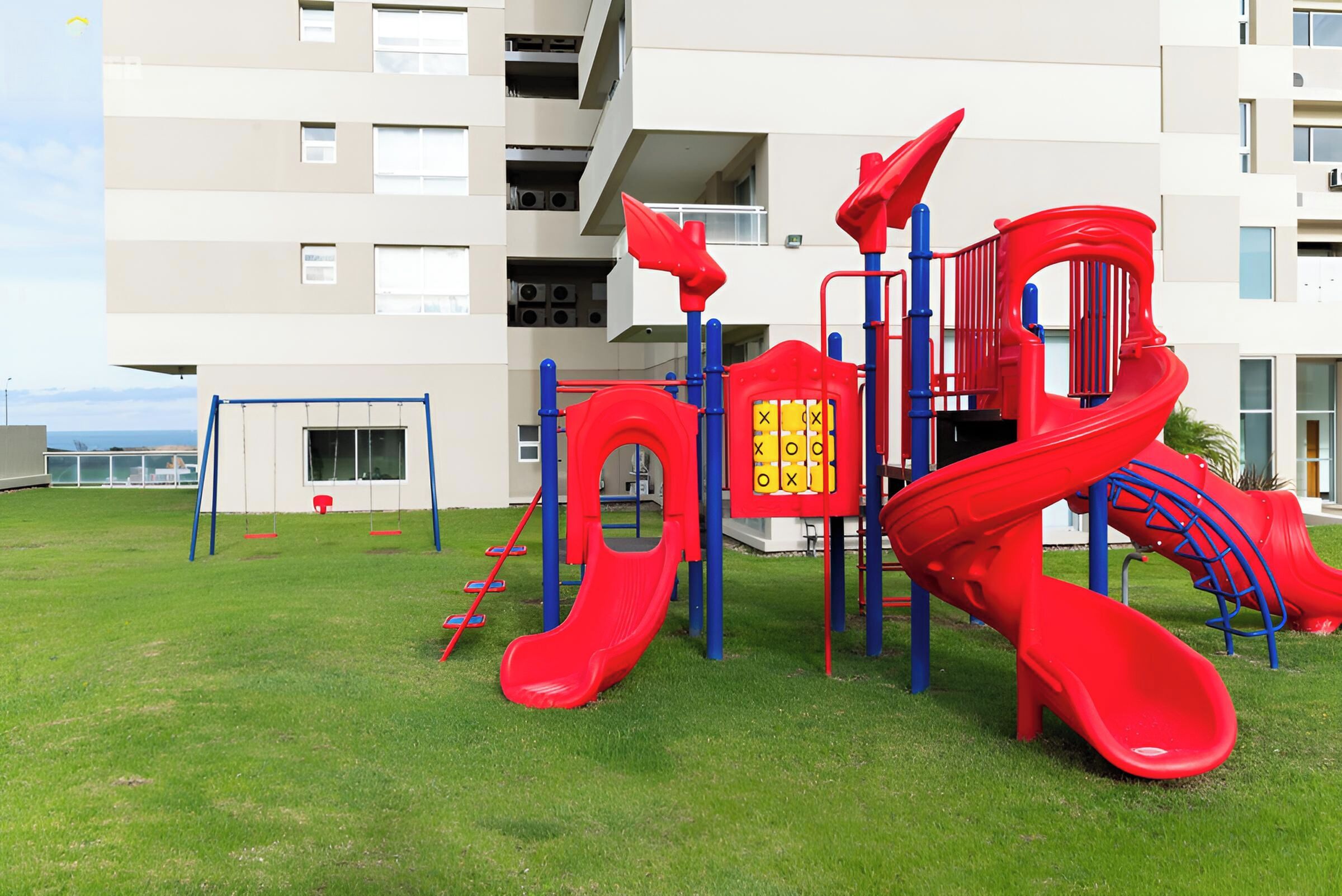 Un parque infantil con un tobogán rojo y estructuras para escalar, ubicado frente a un edificio con múltiples ventanas y balcones.