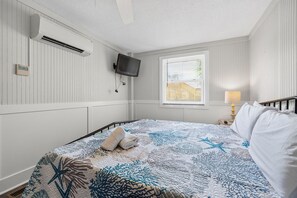 Ein Schlafzimmer mit einem Bett, einem an der Wand befestigten Fernseher, einem Fenster mit Jalousien und einer Nachttischlampe.