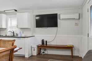 Eine kompakte Küche mit weißen Schränken, einer eingebauten Klimaanlage, einem wandmontierten Flachbildfernseher und einem Essbereich mit einem Holztisch und Stühlen.