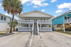 Ein zweigeschossiges Gebäude mit einer zentralen Treppe, einem Balkon im oberen Stockwerk und einem überdachten Bereich, in dessen Nähe ein Auto geparkt ist.