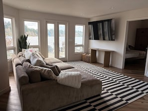 Een woonkamer met een grote bank, een flatscreentelevisie en uitzicht op een waterpartij door grote ramen.