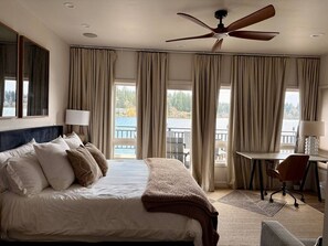 Habitación de hotel con cama, escritorio, silla, ventilador de techo y ventanas grandes con cortinas.