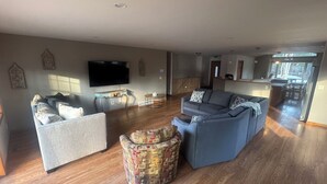 A spacious living room with a large sectional sofa, a patterned ottoman, and a flat-screen TV mounted on the wall. - Timberville - Hank (Gwinn)