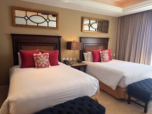 Quarto de hotel com duas camas, cada uma com cabeceira de madeira, mesa de cabeceira com abajur e uma janela grande com cortinas.