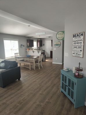 A spacious living area with a dining table and chairs, a sofa, a kitchen area with a sink and cabinets, and a wall with a 'Beach Life' sign.