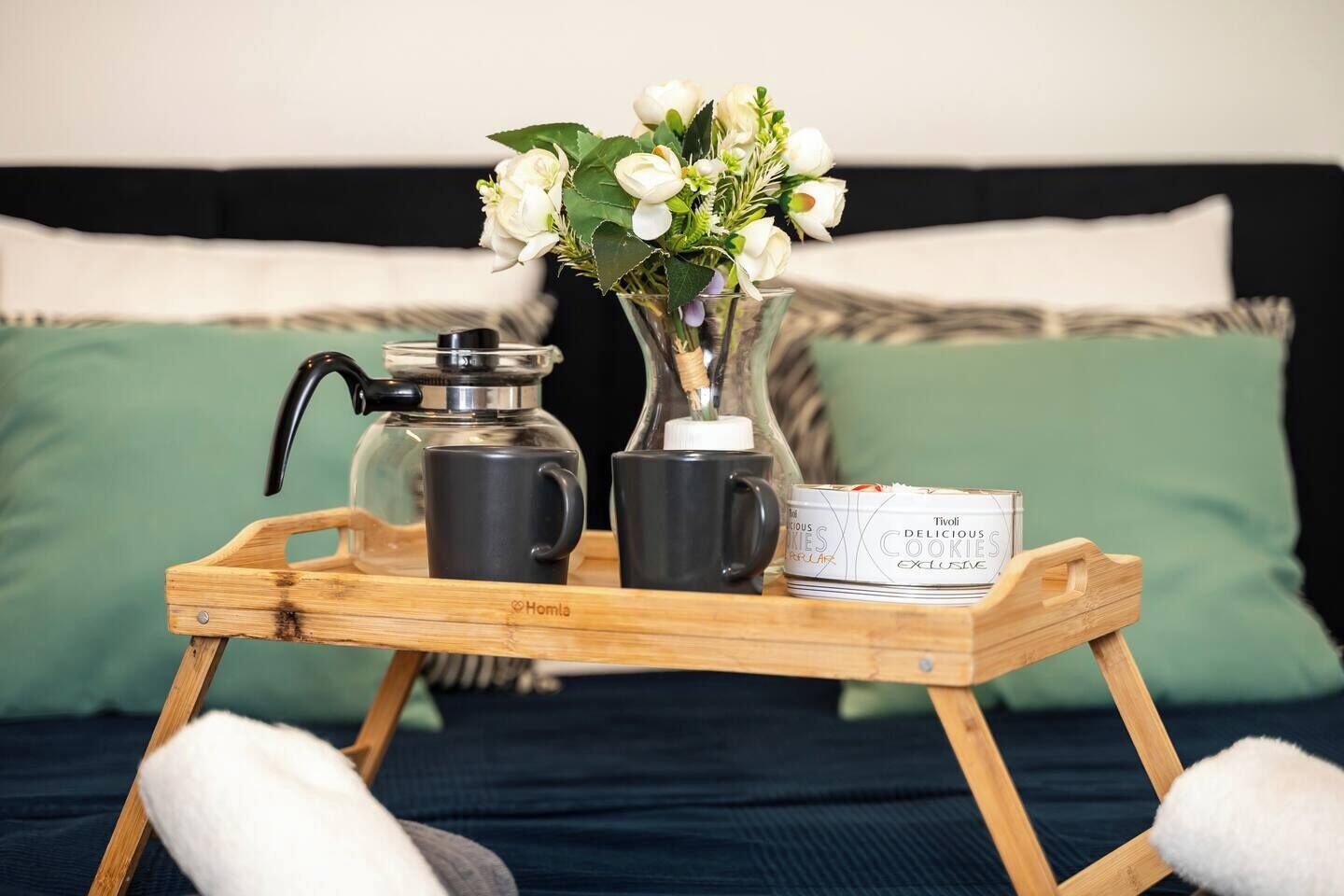 Un plateau en bois avec une machine à café, deux tasses, un vase avec des fleurs et une boîte de biscuits, le tout posé sur un lit avec des oreillers.