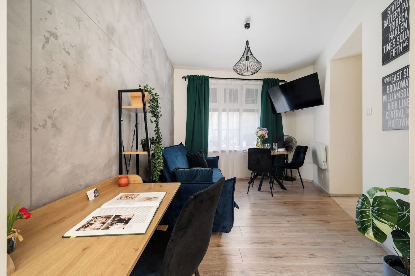 Ett modernt vardagsrum med ett matbord i trä, blå soffor, en tv som är monterad på väggen och ett stort fönster med gröna gardiner.