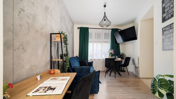 A modern living room with a wooden dining table, blue sofas, a TV mounted on the wall, and a large window with green curtains. - Łódź Płocka Apt – Parks & Public Transport nearby (Łódź)