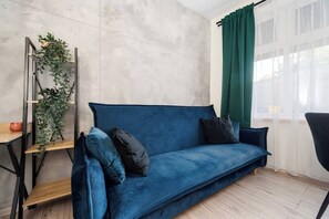 Una sala de estar moderna con un sofá azul, una mesita de madera, una planta verde y vistas a través de cortinas translúcidas.