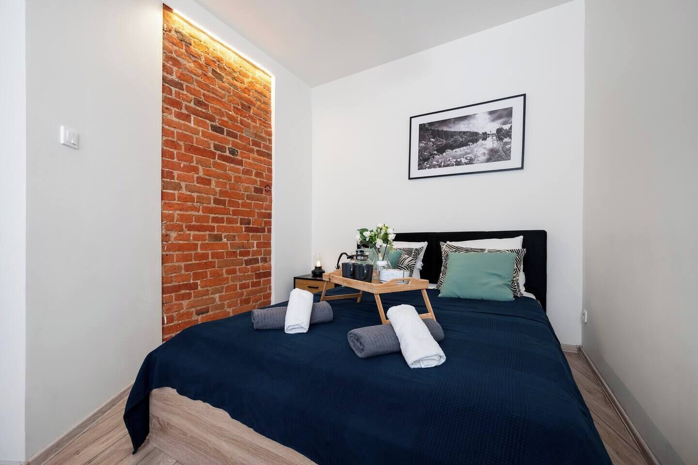 Ett modernt sovrum med en accentvägg i tegel, en snyggt bäddad säng med kuddar och ett brickbord med toalettartiklar, samt en inramad tavla på väggen.