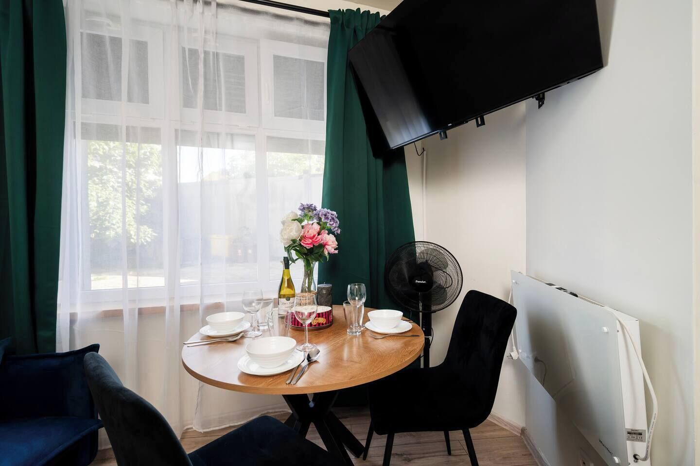 En matplats med ett bord dukat för två personer, en vas med blommor, en fläkt och en tv som är monterad på väggen.
