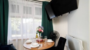 A dining area with a table set for two, a vase with flowers, a fan, and a television mounted on the wall. - Łódź Płocka Apt – Parks & Public Transport nearby (Łódź)