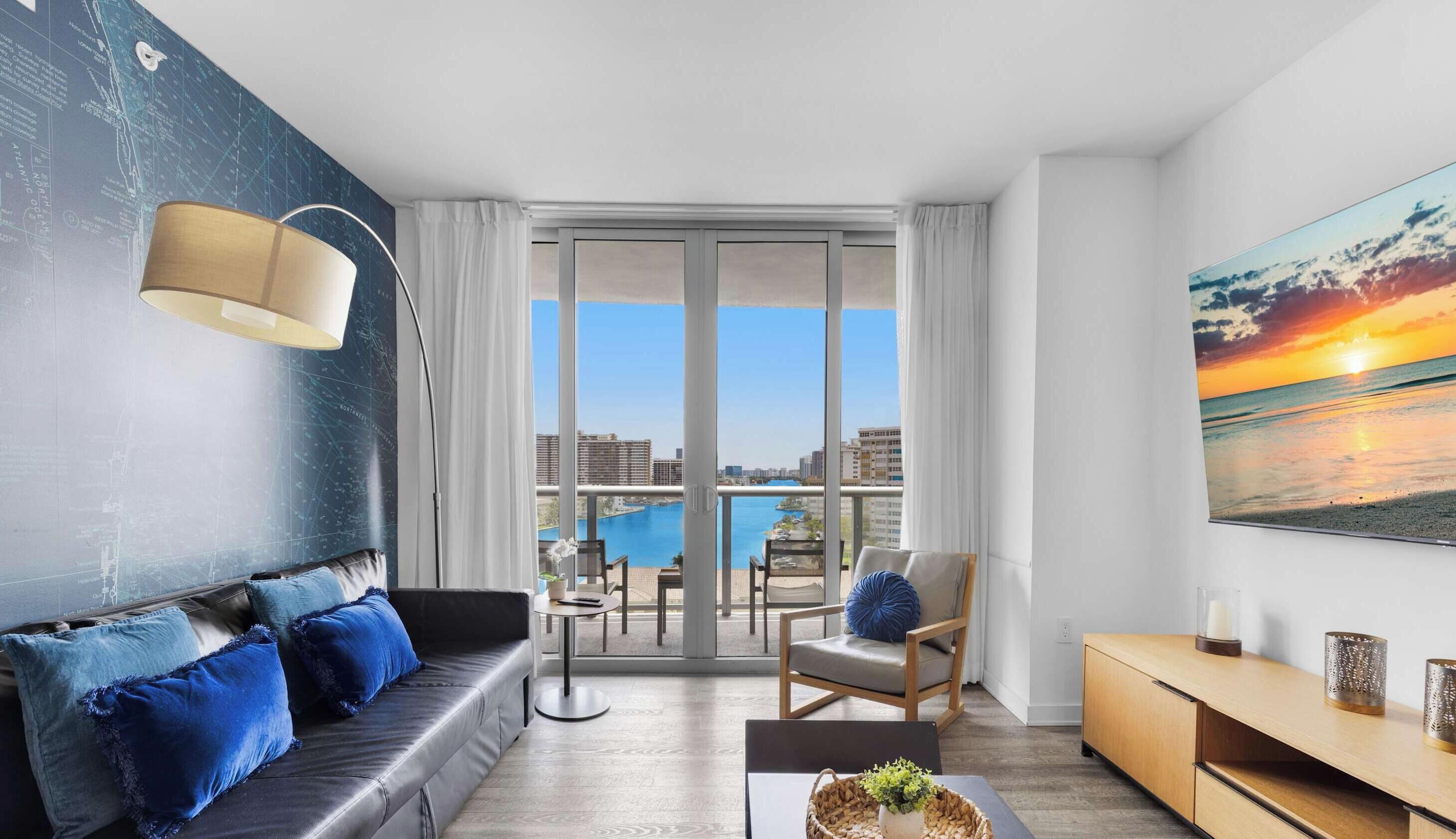 A modern living room with a sofa, a coffee table, and a chair. There is a large window with a view of a city skyline and a body of water.