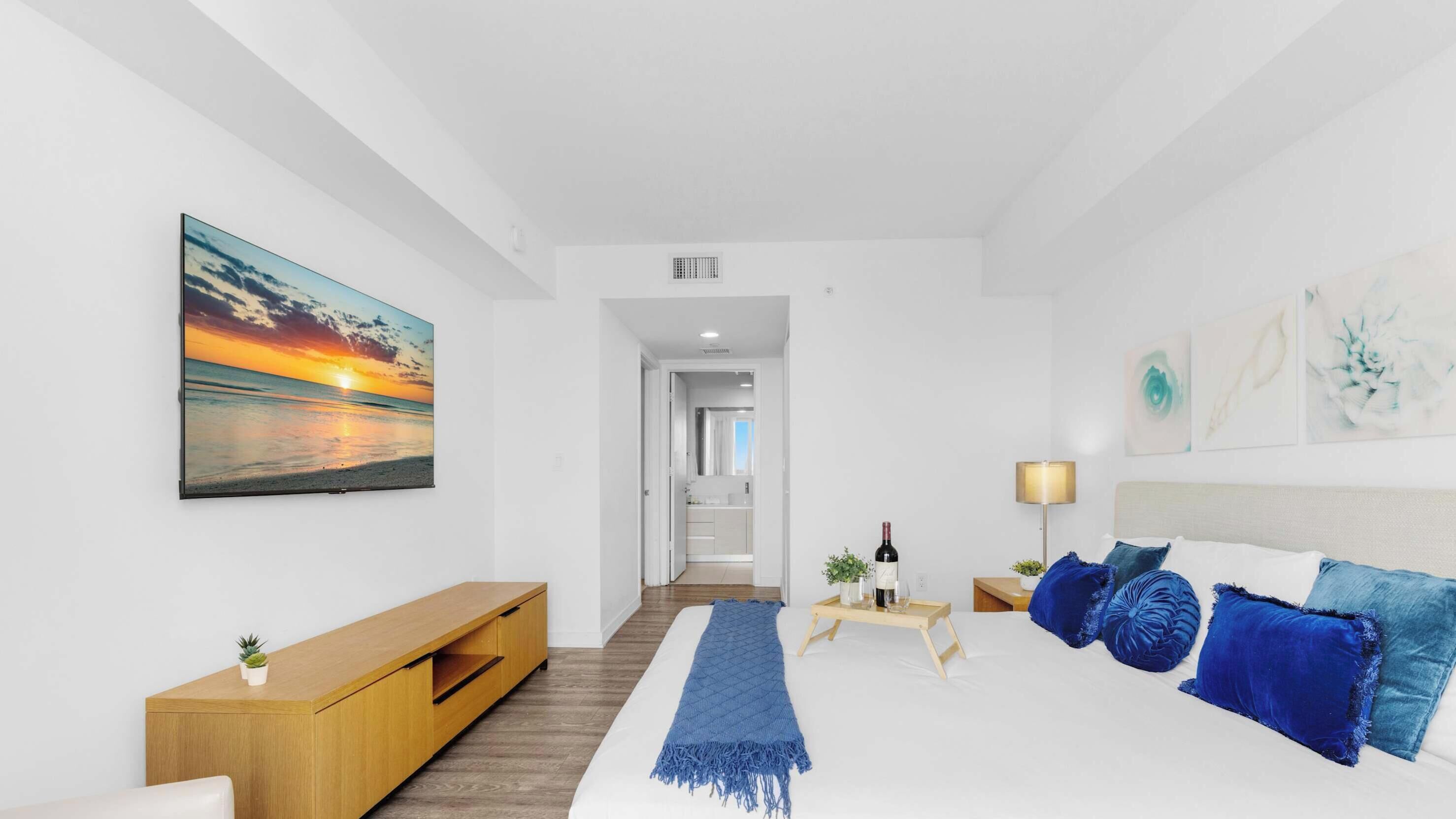 A modern hotel room with a bed, a wooden console table, a painting of a sunset, and a hallway leading to a bathroom.