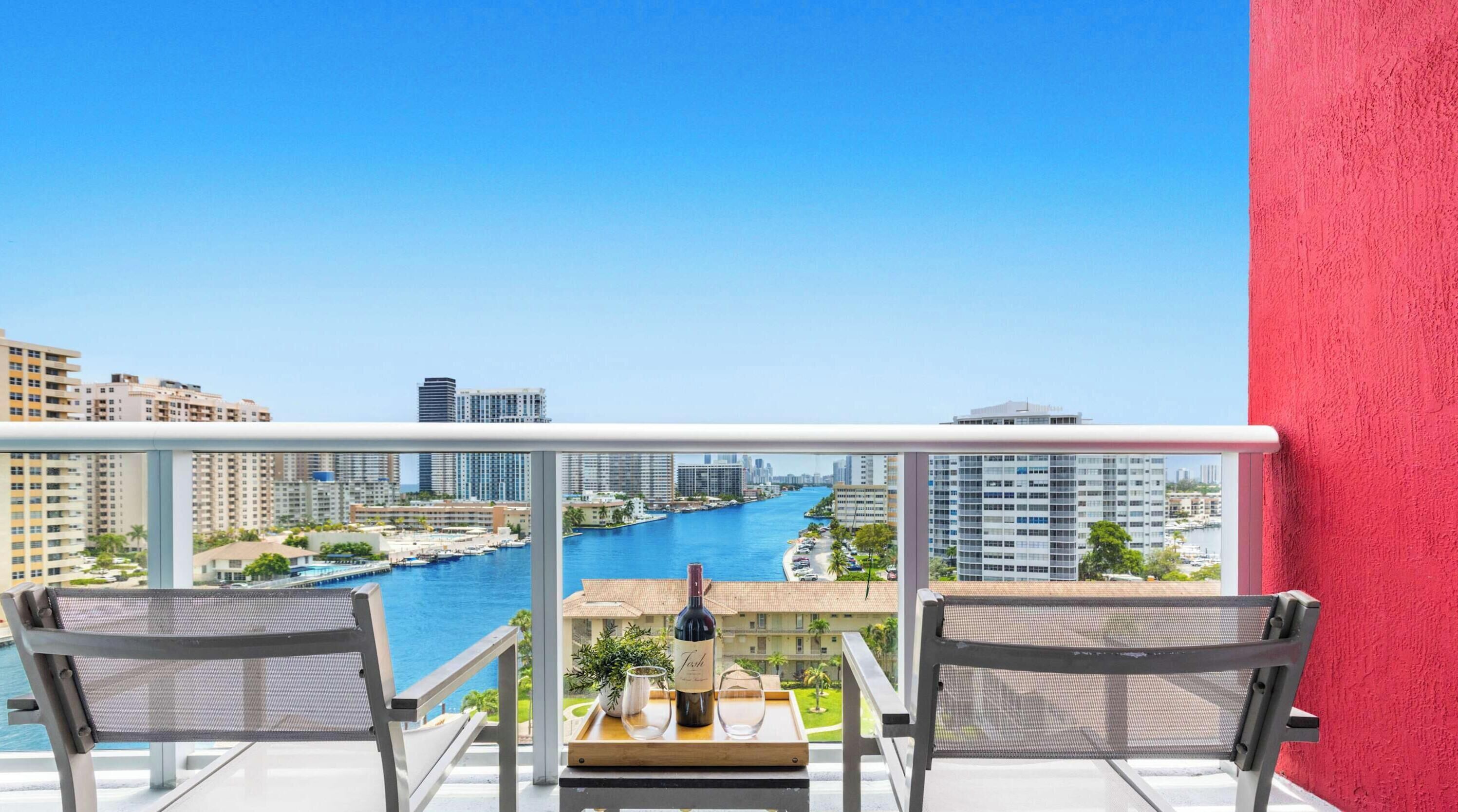 A balcony with two chairs, a table, and a bottle of wine, overlooking a marina and cityscape.
