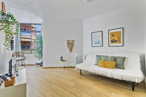 Una sala de estar moderna con un sofá blanco, una mesita auxiliar y una silla. Hay dos cuadros en la pared, una planta en maceta y una ventana con vistas a edificios.