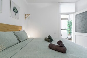 Un dormitorio con cama, cabecero de madera, lámpara de pared y ventana con vista a un edificio.